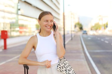 Beautiful stylish young woman is using smartphone, with suitcase outdoors by the airport or railway station waits for taxi cab. Attractive smiling girl ready to travel with luggage in the modern city