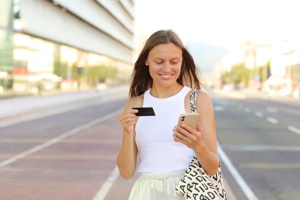 Beautiful smiling young woman consumer with credit card and smartphone looks in camera. people shopping via online application media concept. shopping mall on summer day background with copy space