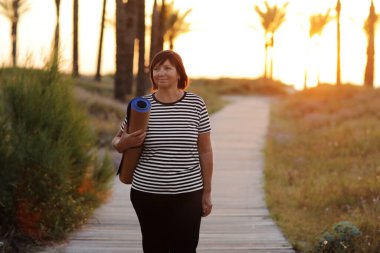 happy middle ages woman with yoga mat and walking to beach to practice yoga, exercising outdoors with happiness feeling on sunrise in the morning. Concept of healthy lifestyle, wellbeing and fitness