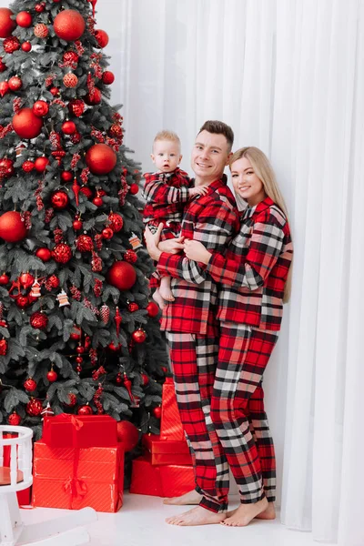 happy young family, mom, dad and little baby boy in the same red pyjamas at home by the stylish decoration Christmas tree are ready to welcome Christmas and New Years. Preparation room to celebration.