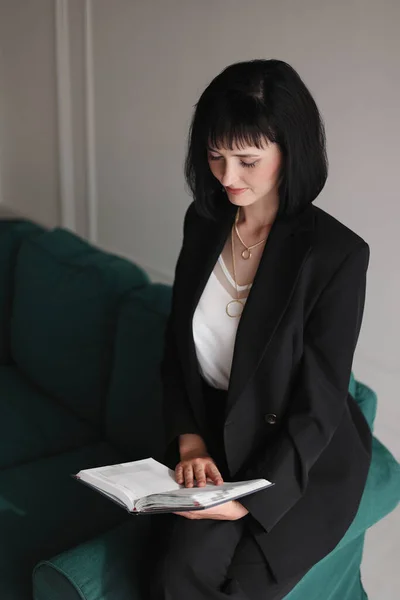 Beautiful business woman writing something in the note pad while sitting on the green sofa at office by window, charming brunette female with short hair in black suit working indoors.