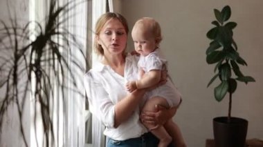 Sevgi dolu genç bir anne kucağında sevimli bir kız bebek kucağında küçük bir çocuğa sarılıyor. Mutlu şefkatli bir anne, evde pencerenin önünde duran küçük çocuğa sarılıyor. Mutlu annelik kavramı.