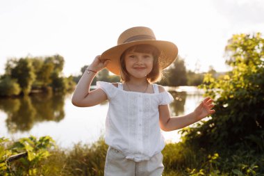 Hasır şapkalı küçük kız yaz tatillerinde eğleniyor. Mutlu çocuk oynuyor ve gölün kenarındaki kameraya bakıyor. Çocuk dışarıda eğleniyor. sağlıklı yaşam tarzı kavramı