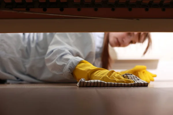 Cleaning and disinfection of surfaces. Woman with yellow rubber gloves and rag cleaning floor under bed. Beautiful woman cleaning house