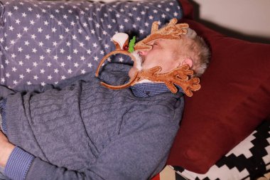 50-60 yaşlarında bir adam kafasında geyik boynuzu şeklinde süslerle kanepede uyuyor. Noel ve Yeni Yıl partisinden sonra dinlenen adam.