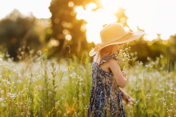 smiling child girl in a big mommys straw hat with bouquet of wildflowers in a green grassy meadow on summer sunny day. Happy childhood concept. Copy space