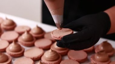 Making french desert macarons. Chef hands in black gloves with confectionery bag add cream to the ready halves of brown chocolate macaroons at pastry shop. cooking, food and baking concept