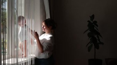 happy young mother with baby girl or boy resting at home near big window. Infant daughter or son is sitting on windowsill and looking out through window with mom. family. Maternity concept.Motherhood