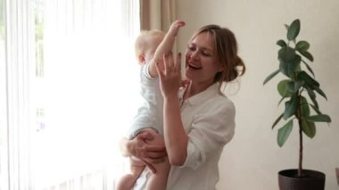 Young smiling mother caressing and having fun with her newborn baby kept by mom in arms by window at home in the morning. Concept of family, fatherhood, motherhood, parenthood, childhood, life.