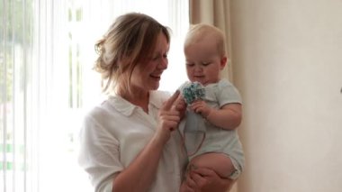 Young smiling mother caressing and having fun with her newborn baby kept by mom in arms by window at home in the morning. Concept of family, fatherhood, motherhood, parenthood, childhood, life.