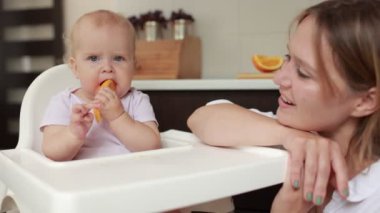 Adorable baby girl eating orange. Little daughter sitting in feeding chair and eating slice of orange at kitchen and playing with mom. Juicy fruits oranges are for a healthy diet. Happy childhood