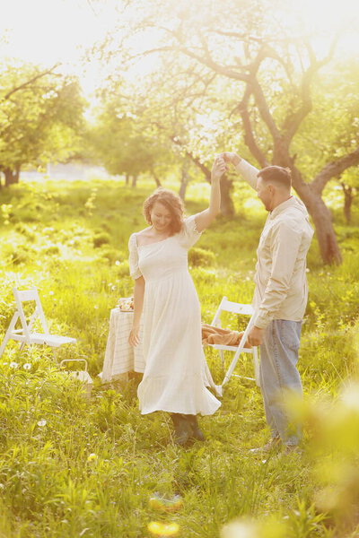 Молодая пара, наслаждающаяся едой и напитками в красивом летнем зеленом парке на романтическом пикнике, красивые мужчина и женщина с кудрявыми волосами обнимаются на открытом воздухе.