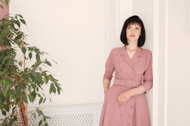 Çekici, gülümseyen esmer ve kısa saçlı bir kadının portresi. Beyaz, şık bir duvar arkasında yeşil çiçekli yalnız kalıyor. Yumuşak pembe elbiseli kadın kameraya bakıyor..