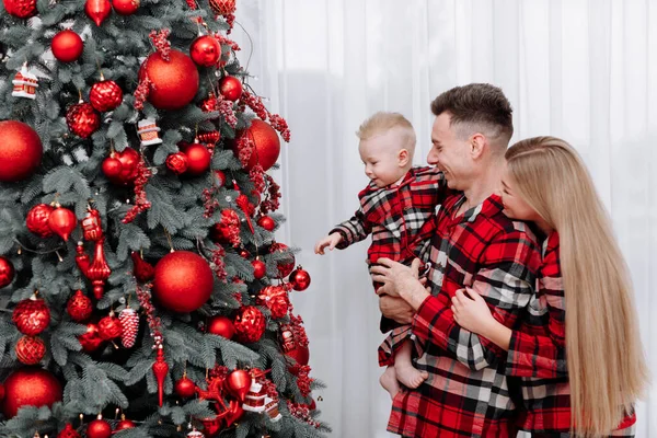 happy young family, mom, dad and little baby boy in the same red pyjamas at home by the stylish decoration Christmas tree are ready to welcome Christmas and New Years. Preparation room to celebration.