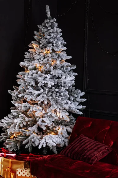 Stylish dark Christmas living room interior with red velvet sofa, white christmas tree with garland lighting and presents, gift boxes with ribbons under it. Family time. Marry Christmas and New Year.