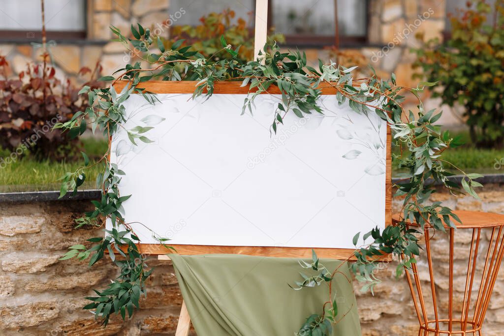 Wedding wooden board, mockup, invitation easel, with white space for an inscription, decorated with fresh greenery and green cloth. Wedding frame outdoors. Festive decor, copy space