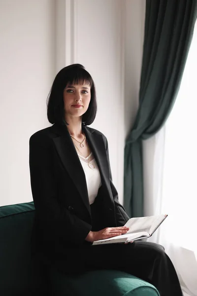 Beautiful business woman writing something in the note pad while sitting on the green sofa at office by window, charming brunette female with short hair in black suit working indoors.