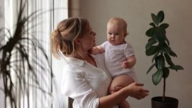 Sevgi dolu genç bir anne kucağında sevimli bir kız bebek kucağında küçük bir çocuğa sarılıyor. Mutlu şefkatli bir anne, evde pencerenin önünde duran küçük çocuğa sarılıyor. Mutlu annelik kavramı.