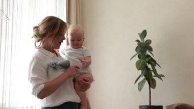 Young smiling mother caressing and having fun with her newborn baby kept by mom in arms by window at home in the morning. Concept of family, fatherhood, motherhood, parenthood, childhood, life.