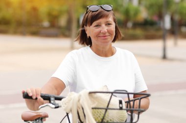 Güneşli bir günde bisiklet süren aktif orta yaşlı kadın portresi. Beyaz tişörtlü olgun bayan parkta bisiklet sürüyor. Boş zaman ve yaşam tarzı konsepti, gülen kadın yazın bisikletiyle bisiklet sürüyor..