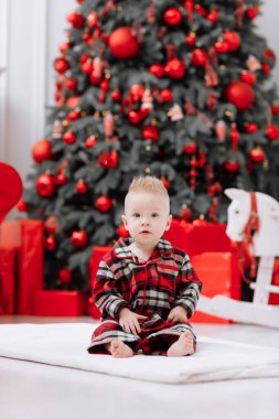 Adorable caucasian baby boy is sitting and holding gift toy Santa Claus near a Christmas tree, many festive gift boxed and red sofa at home, baby infant. Xmas and New Year celebrations concept