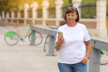 Beautiful mature woman holding smartphone and smiling while standing on the street near bicycles. Female with mobile phone in her hand using bike rental service on summer sunny day. Active lifestyle.