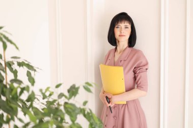 Zarif, yumuşak, pembe elbiseli çekici genç kadın elinde sarı klasör dolusu evrak işi var. Sahte alanla kameraya bakıyor ve ev bitkileriyle ofiste duruyor. Eğitim, iş, yaşam tarzı.