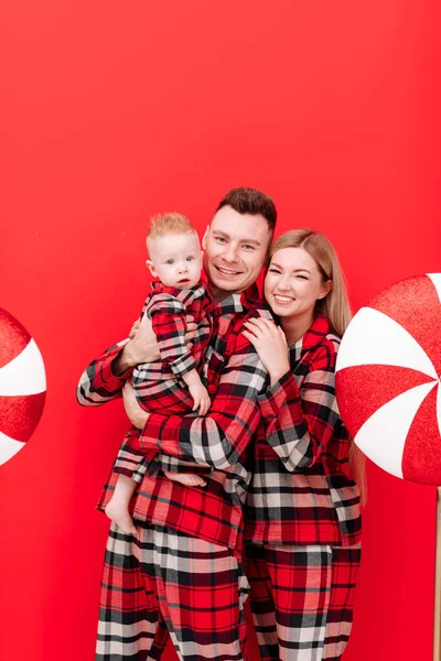 Young dad, mom and little son in red plaid pyjamas on red Christmas background with big lollipop. Amazing happy family looking in camera. Happy mothers day, fathers day. Parenthood. Holiday concept.