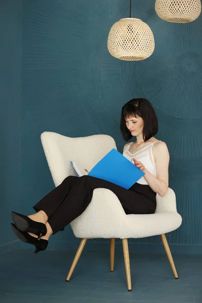 Young self confident woman with short hair sitting, relaxing on chair and holding blue folder and checking documents. Successful brunette female. Education, business, lifestyle concept.
