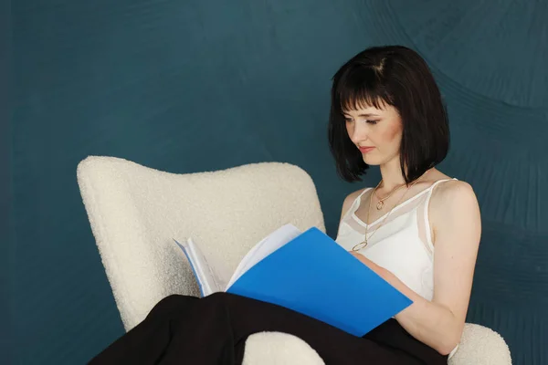 Young self confident woman with short hair sitting, relaxing on chair and holding blue folder and checking documents. Successful brunette female. Education, business, lifestyle concept.