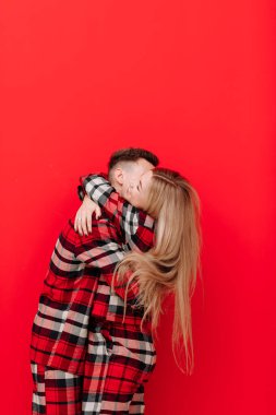 Happy Saint Valentine's Day. Young couple in the same pyjamas is kissing and hugging on red background.Man and woman in love are enjoying spending time together while celebrating, copy space for text.