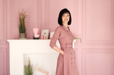 Çekici, gülümseyen esmer ve kısa saçlı kadının portresi kollarını kavuşturmuş ve iç mekanda tek başına kalıyordu. Yumuşak pembe elbiseli kadın kameraya bakıyor..