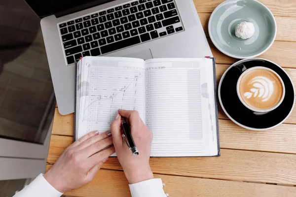 flat lay. cropped photo of business woman in cafe is taking notes and drawing graphics and charts in paper notebook. On table is laptop, sweets and a cup of coffee. Student learning finance online