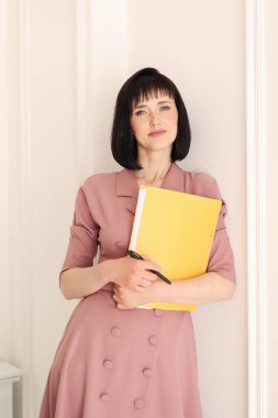 Zarif, yumuşak, pembe elbiseli çekici genç kadın elinde sarı klasör dolusu evrak işi var. Sahte alanla kameraya bakıyor ve ev bitkileriyle ofiste duruyor. Eğitim, iş, yaşam tarzı.