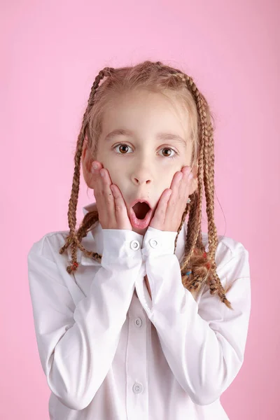 Surprised, wow, shock. Portrait of cute girl with funny hairdo pigtails in white shirt and hands near open mouth on pink background. Happy childhood concept. Emotions, facial expression concept.