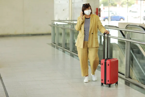 在机场 戴着防毒面具的成年女性正在用智能手机交谈 身穿黄色西服 头戴红色行李箱 戴着防护卫生面罩等待飞行的女人 复制空间 — 图库照片