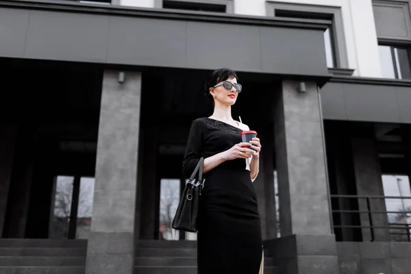 Smiling business woman in sunglasses walks down the business central city street with offices and holding cup of coffee and has break. Pretty woman in has a business dress code and wearing in dress.