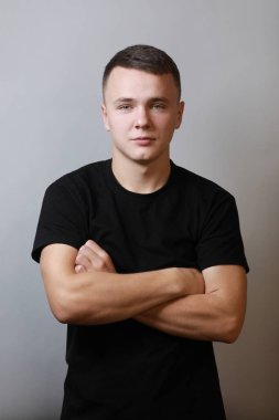 Kendine güvenen esmer, yakışıklı, siyah tişörtlü, göğsünde çapraz ellerle gri arka planda kameraya bakan genç adamın yakın portresi.
