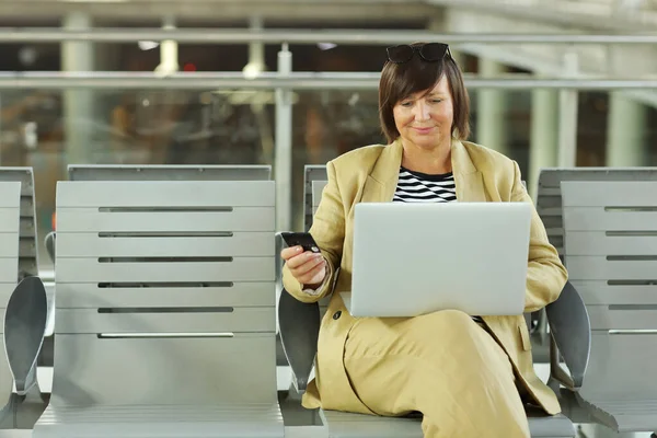 Smiling middle ages woman using credit card and laptop computer making online payment while checking flight or online check-in at airport. Mature woman holding debit card for internet banking account.