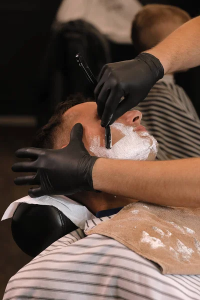 Barber Man is shaving client with a vintage straight razor in a barbershop. Shaving cream, foam. Classic shave by stainless steel straight edge razor