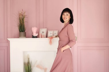 Çekici, gülümseyen esmer ve kısa saçlı kadının portresi kollarını kavuşturmuş ve iç mekanda tek başına kalıyordu. Yumuşak pembe elbiseli kadın kameraya bakıyor..