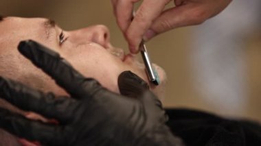 Koruyucu eldivenli bir berber, jiletle genç bir adamı tıraş eder. Bir berber dükkanında ustabaşı bir adamın bıyığını ve sakalını bıçakla kesiyor. Şık bir berber dükkanı..