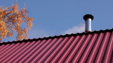 Özel bir evin çatısı bacası, bacadan çıkan beyaz duman, sonbaharda evin serinletilmesi ve ısıtılması. Parlak mavi gökyüzü ve sarı sonbahar ağacı dalları.