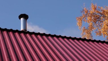 Bir binanın kırmızı çatısındaki bacadan çıkan duman mavi gökyüzüne ve bir sonbahar ağacının dallarına karşı.