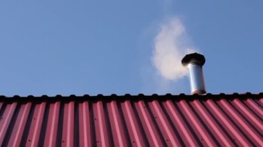 Bir evin çatısındaki bacadan duman, güzel mavi gökyüzüne karşı kırmızı metal bir çatı, bir ev ısıtma ve konfor kavramı..