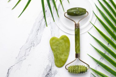 Evde yüz bakımına hazır. Guasha ve yeşil taştan yapılmış yüz masajı silindiri. Yeşil tropikal yapraklı beyaz mermer arka planda kozmetik kaldırma kiti.