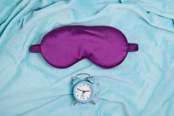 トルコ石の上に小さな青い目覚まし時計を持つ紫の睡眠マスクが背景を果たした 良い睡眠のための快適なサテンマスク — ストック写真