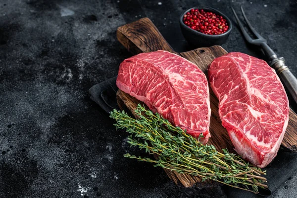 Fresh Raw Top Blade or flat iron beef meat steaks on a butcher cutting board. Black background. Top View. Copy space