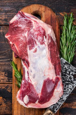 Tahta bir kasapta çiğ kuzu eti ya da keçi omuzu eti, satırla tahtayı keser. Koyu ahşap arka plan. Üst görünüm