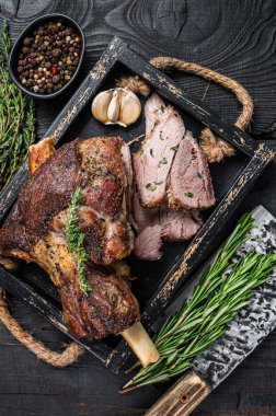 Ahşap bir tepside et satırıyla dilimlenmiş kızarmış koyun eti bacağı. Siyah ahşap arka plan. Üst görünüm